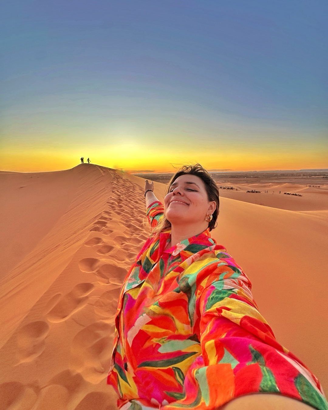 Una persona tomándose un selfie con una camisa colorida en un paisaje desértico con dunas de arena y cielo despejado.