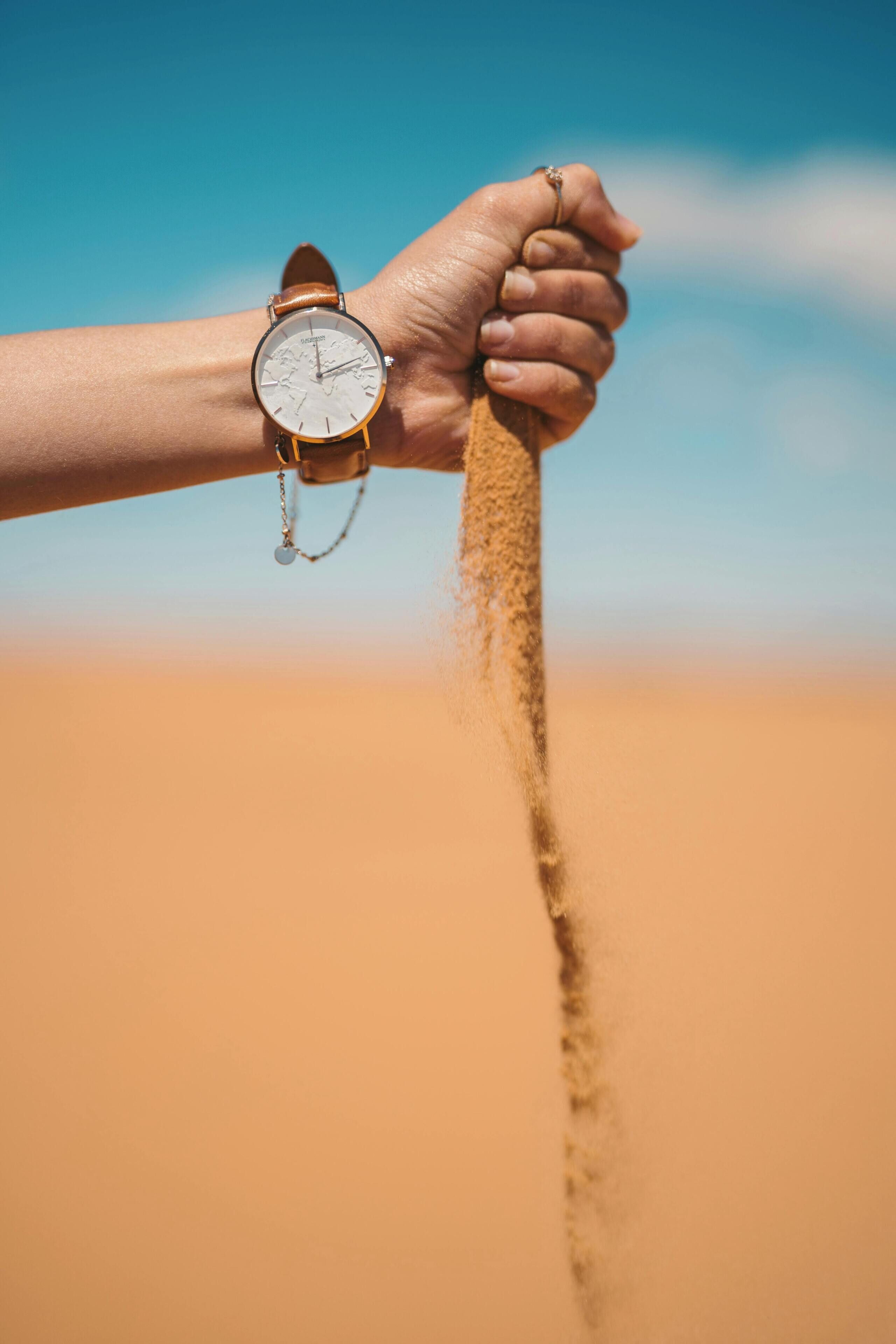 A hand holding a watch with a leather strap and a chain, against a desert background.