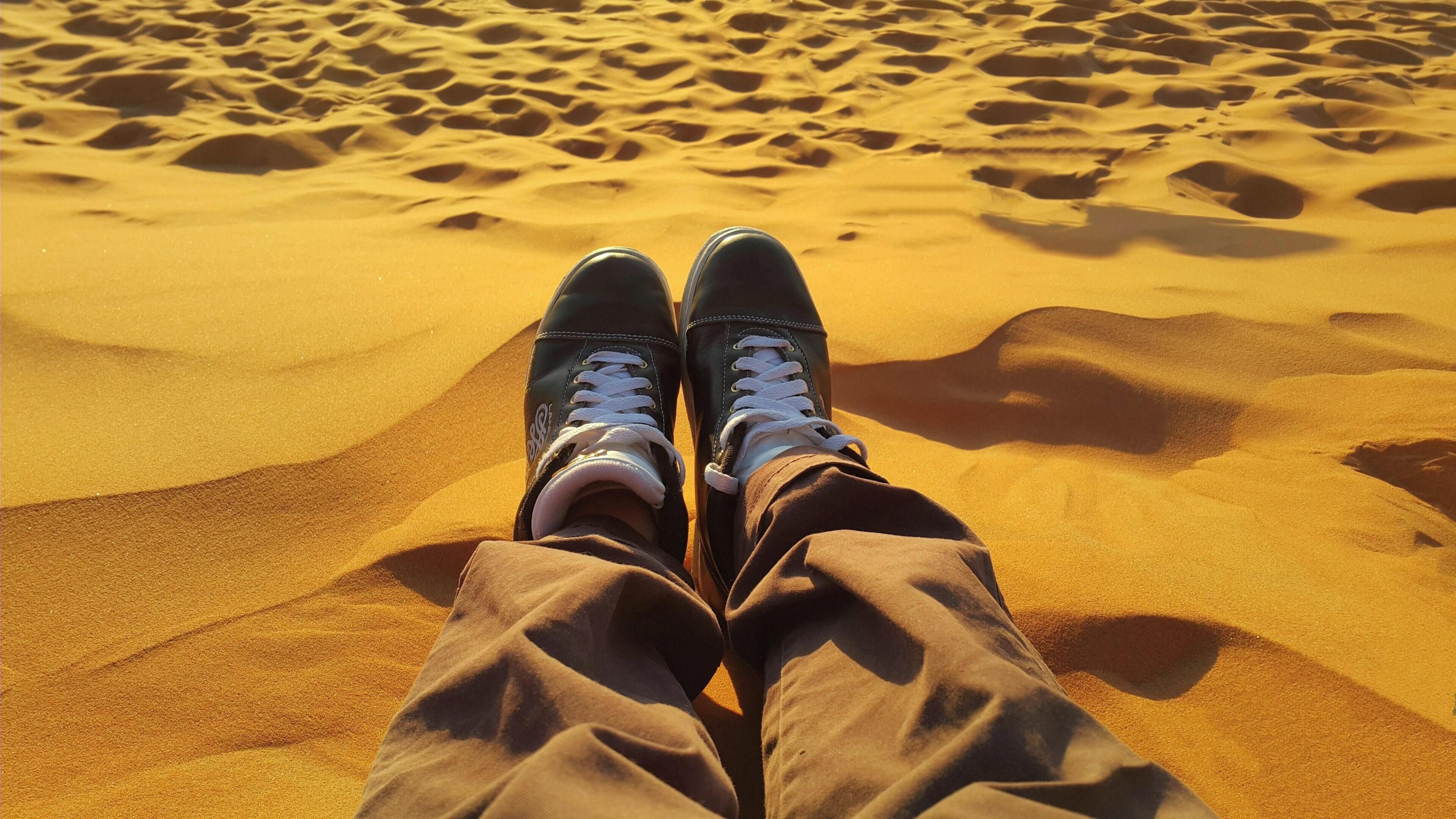 A pair of feet in brown pants and black shoes with white laces, resting on a sandy surface with undulating patterns.