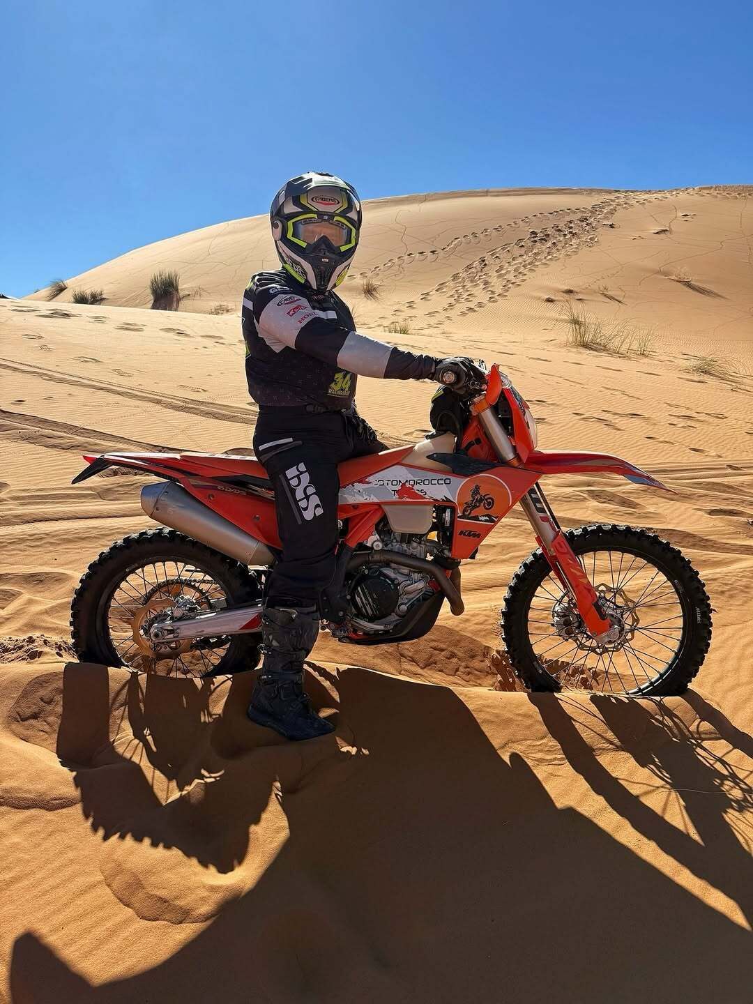 Una persona conduce una moto de tierra roja en un desierto con dunas de arena al fondo.