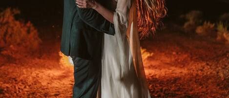A couple embracing in a dimly lit outdoor setting with a ground covered in dry leaves.