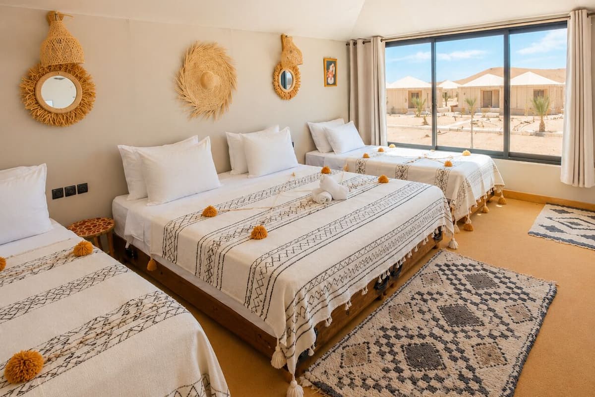 Habitación de hotel con tres camas, un ventanal grande y vistas a tiendas de campaña en el desierto.