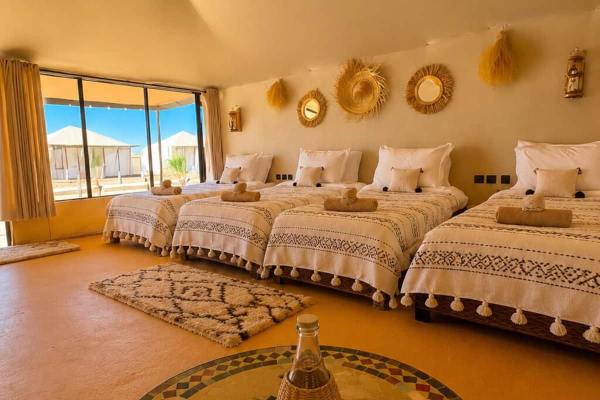 Una habitación con una cama grande, una mesa redonda con una botella y vasos, y vistas a tiendas de campaña desde la ventana.