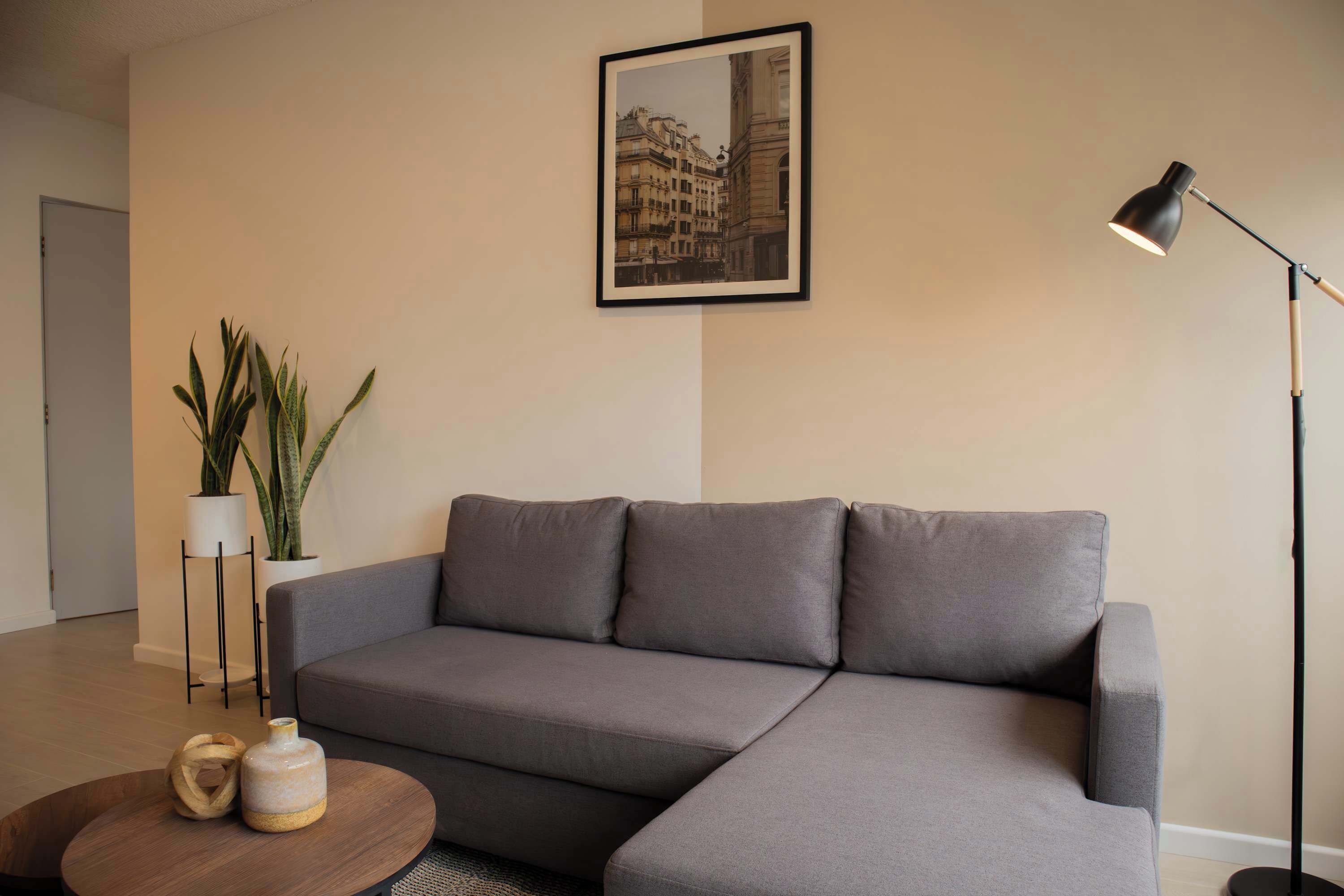 Ein modernes Wohnzimmer mit einem grauen Ecksofa, einem runden Couchtisch aus Holz, einer Stehlampe, einem Bilderrahmen mit Stadtansicht und zwei Kübelpflanzen.