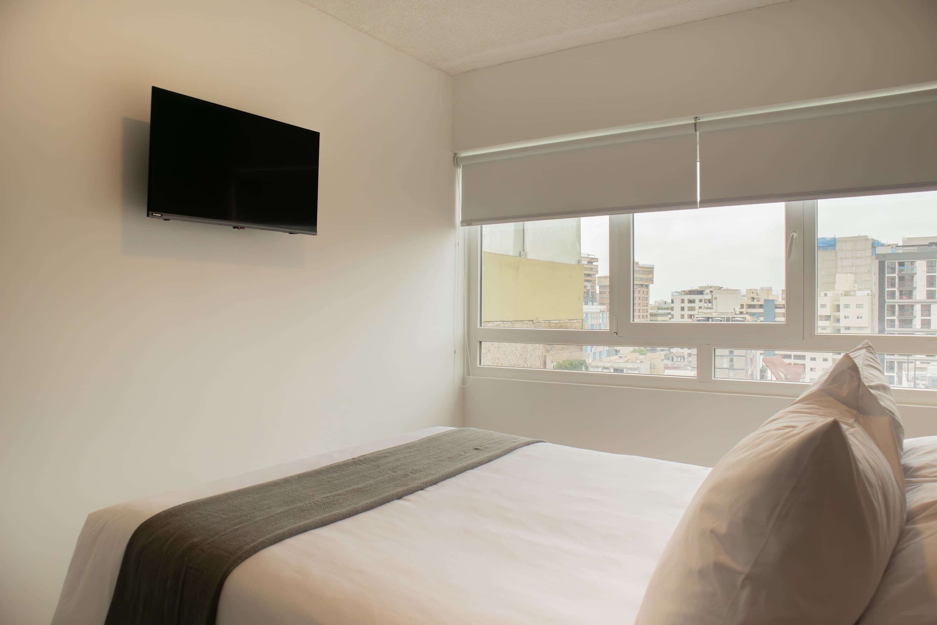 Habitación de hotel con cama, televisor de pantalla plana en la pared y ventana con vistas a edificios.
