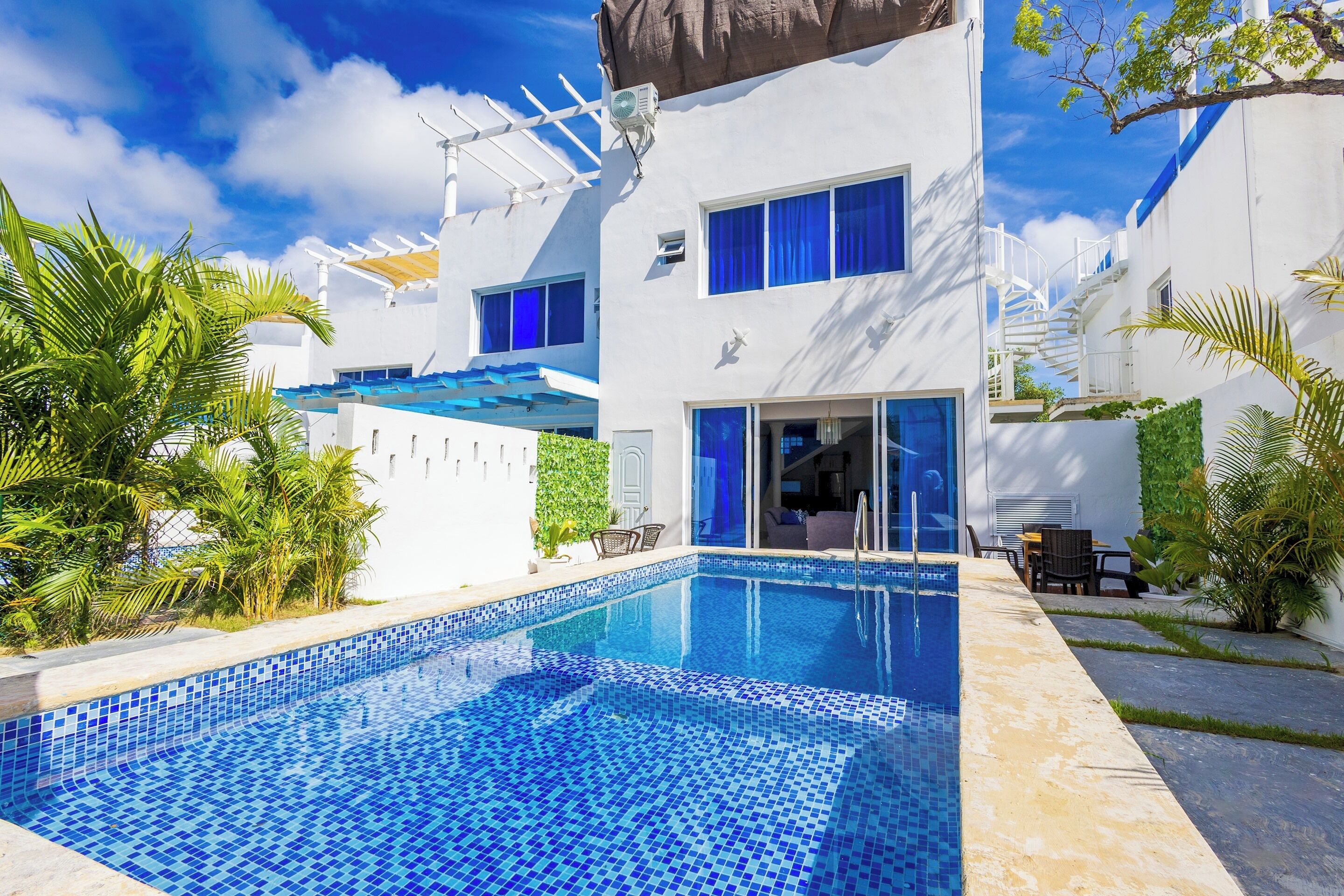 A swimming pool with blue mosaic tiles, surrounded by white buildings with blue accents, palm trees, and a clear sky.