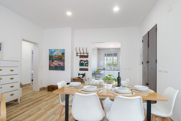 A dining area with a wooden table set for four, white chairs, a kitchen in the background, and a painting on the wall.