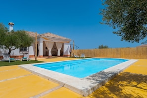 A swimming pool with a tiled deck, a covered outdoor seating area, and a wooden fence.