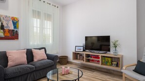 Un salon moderne avec un canapé gris, une table basse en verre, un téléviseur à écran plat, une bibliothèque et une fenêtre ornée de rideaux légers.