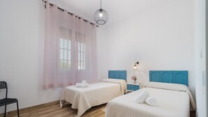 Une chambre avec deux lits, une chaise, une table de chevet avec une lampe et une fenêtre avec des rideaux légers.