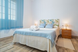 A neatly made bed with a blue bedspread, two pillows, and rolled towels. There is a wooden nightstand with a lamp, a wall-mounted painting, and a window with blue curtains.