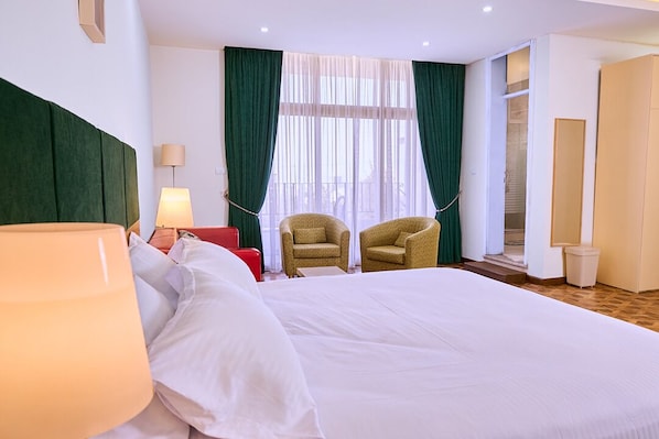 Habitación de hotel con una cama grande, dos sillones, una lámpara y vistas al exterior a través de cortinas translúcidas.