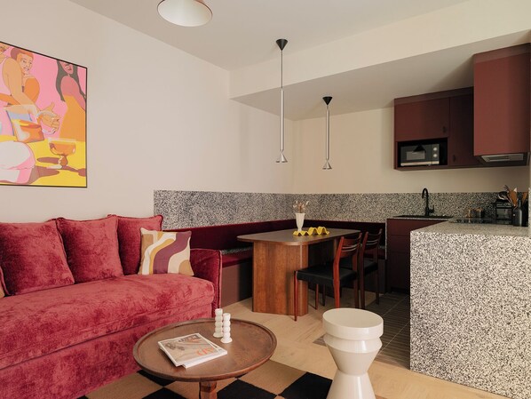 A modern living area with a red sofa, a coffee table with a magazine, a small dining table with chairs, and a kitchen with a microwave and cabinets.
