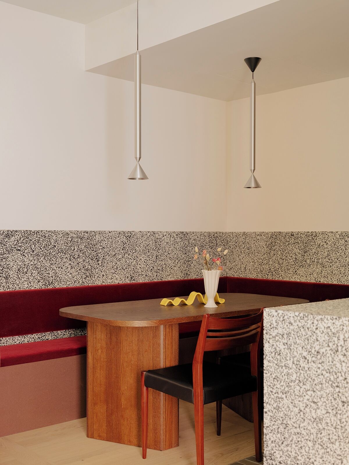 Een eethoek met een houten tafel, een rode met kussens beklede bank en een witte vaas met bloemen.