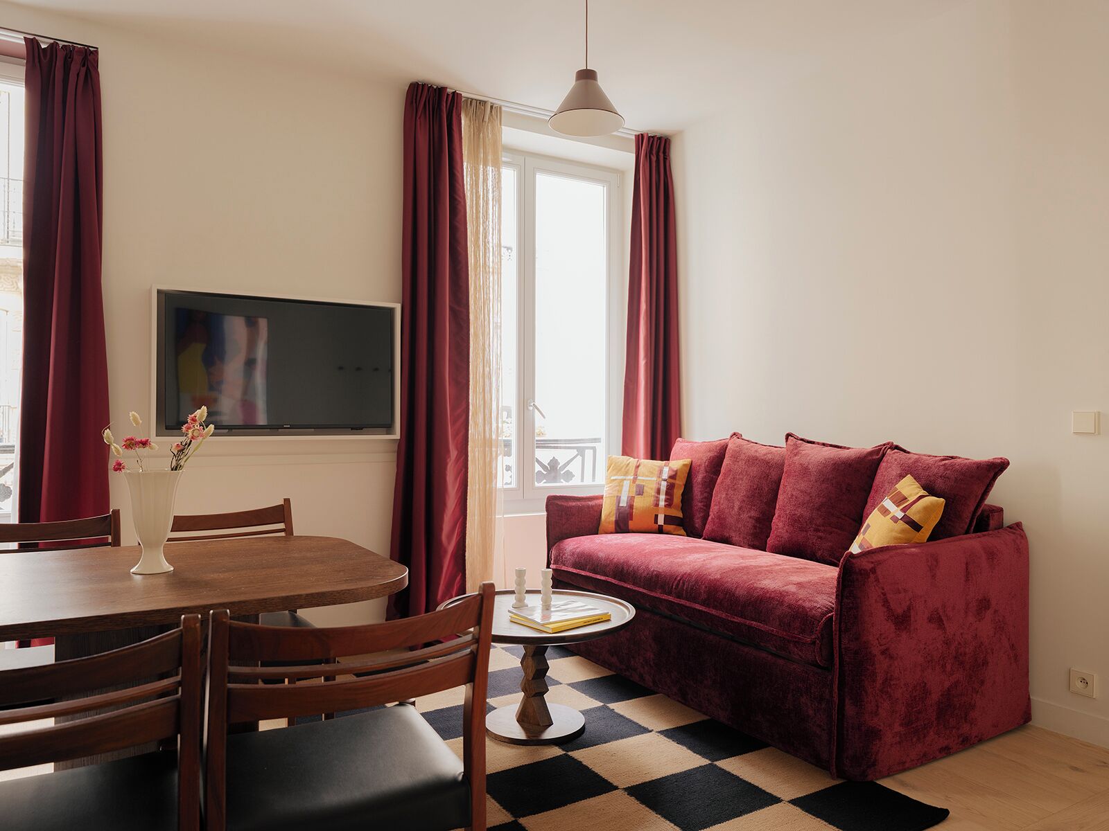 A room with a round dining table, wooden chairs, a red sofa with patterned cushions, a small side table, and a TV mounted on the wall.