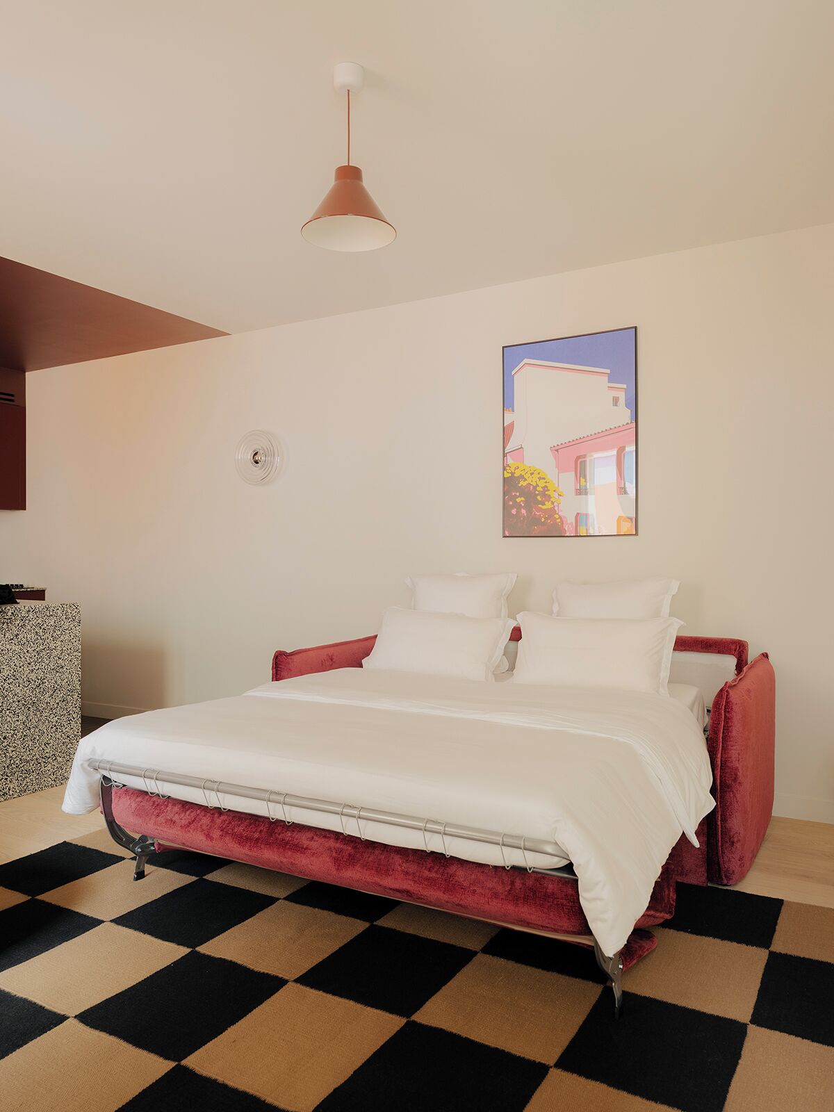 Une chambre avec un sol à carreaux, un lit avec une literie blanche et une tête de lit rouge, un luminaire suspendu et un tableau accroché au mur.