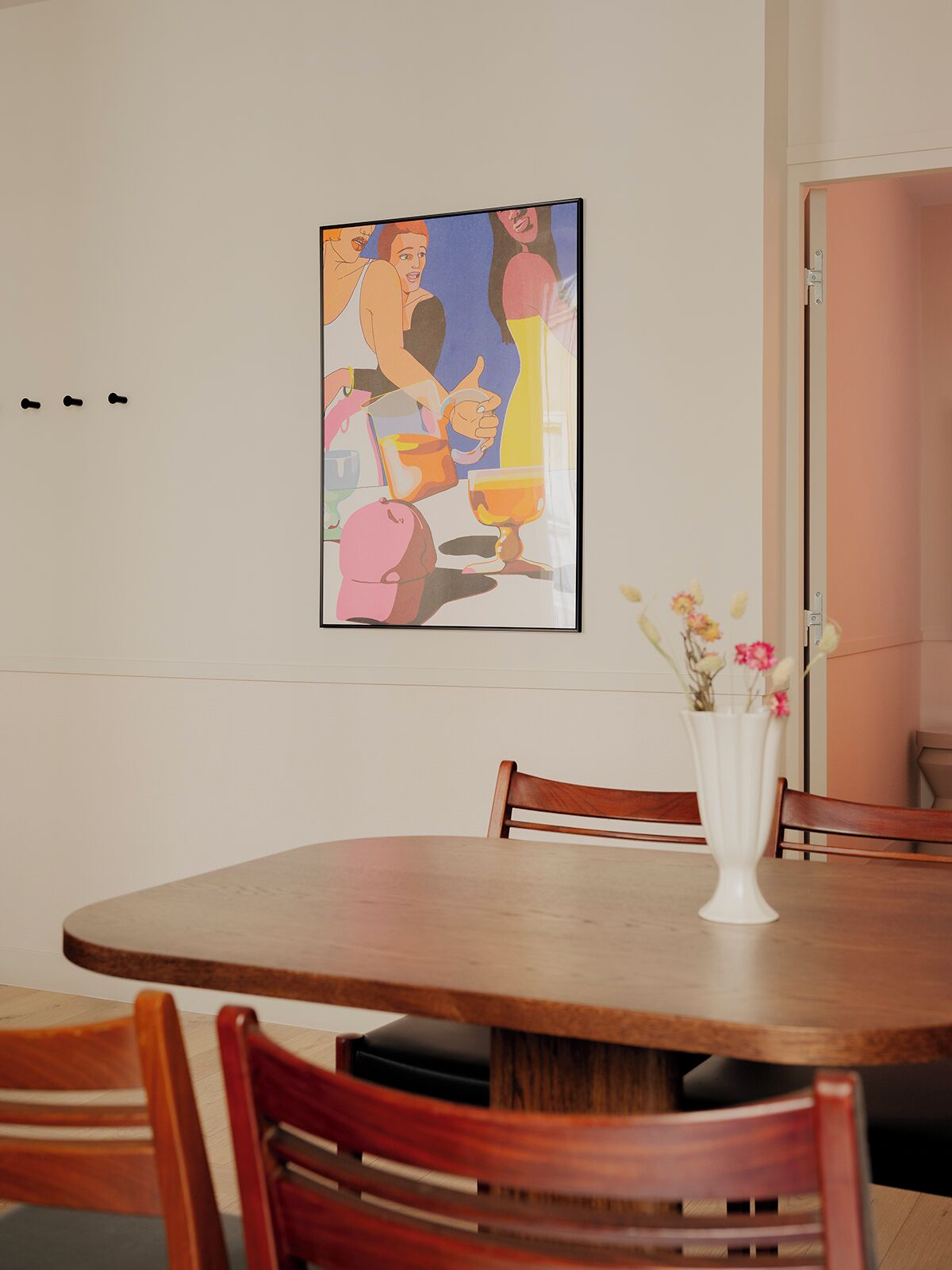 A dining area with a round wooden table, a white vase with flowers, and a colorful abstract painting on the wall.