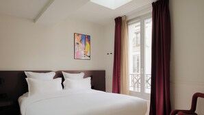 A hotel room with a large bed, a nightstand, a chair, a window with curtains, and a painting on the wall. - Edgar Suites - Lemercier  (Paris)