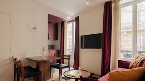 A compact living space with a dining area, a sofa, a TV, and a window with curtains. - Edgar Suites - Lemercier  (Paris)