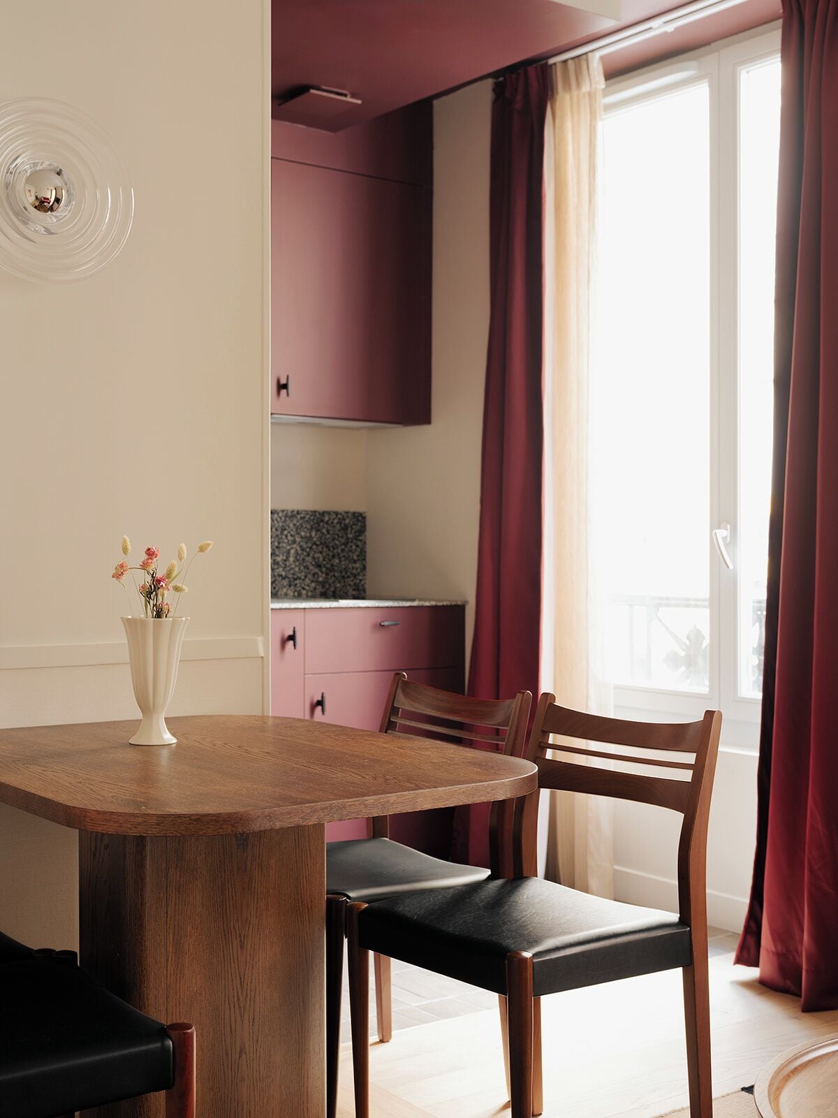 Een eethoek met een ronde houten tafel, twee stoelen, een vaas met bloemen, en een keuken op de achtergrond.