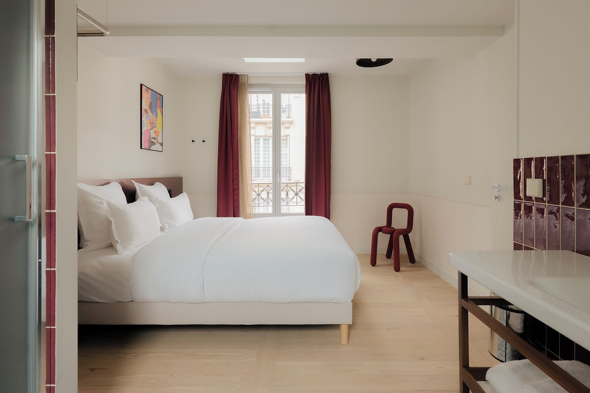 Un dormitorio con cama, una ventana con cortinas, un taburete pequeño y un baño con espejo y azulejos.