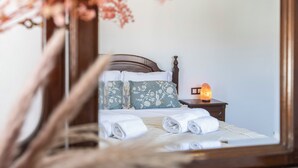Un lit bien fait, agrémenté d’un coussin à motifs floraux, d’une serviette blanche et d’une table de chevet sur laquelle brûle une bougie.