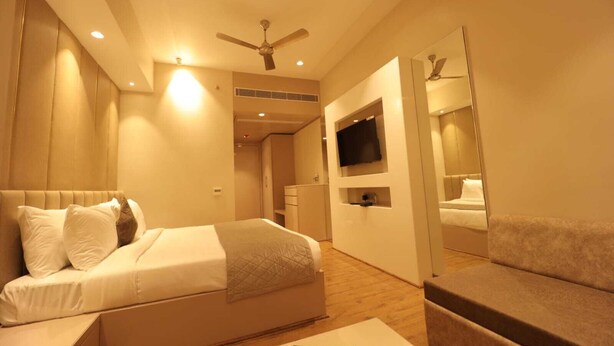 A modern hotel room with a large bed, a sofa, a TV mounted on the wall, and a ceiling fan. - MVR Grand Hotel (Kanpur)