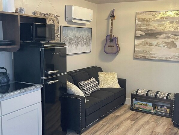 A compact living space with a kitchenette, a black sofa with patterned cushions, a guitar, and a framed picture on the wall.