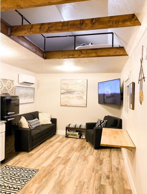 A loft-style living room with exposed wooden beams, a black sofa, a wooden coffee table, and a flat-screen TV mounted on the wall.
