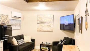Una sala de estar tipo loft con vigas de madera a la vista, un sofá negro, una mesa de centro de madera y un televisor de pantalla plana montado en la pared.