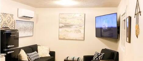 A loft-style living room with exposed wooden beams, a black sofa, a wooden coffee table, and a flat-screen TV mounted on the wall.