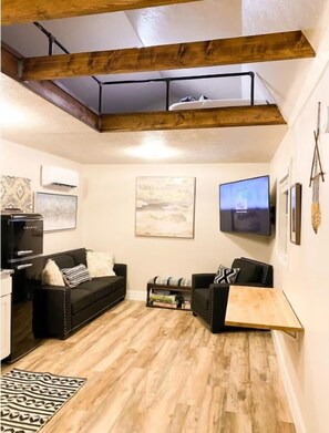A loft-style living room with exposed wooden beams, a black sofa, a wooden coffee table, and a flat-screen TV mounted on the wall.