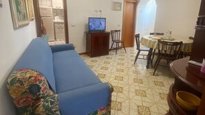 A living room with a blue sofa, a television, a dining table with chairs, and a kitchen area in the background. - CASA DONNA CONCETTA (Positano)