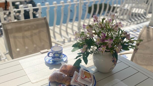 A table set with a floral centerpiece, croissants, pastries, and a blue and white tea set on a terrace overlooking a coastal landscape. - CASA DONNA CONCETTA (Positano)