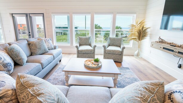 A spacious living room with a large window, a sofa, two armchairs, a coffee table, and a TV on the wall. - New Construction! Dogs OK, Elevator, Direct Beach Access! (Eastpoint)
