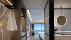 Una habitación de hotel moderna con una cama grande, un baño con un espejo amplio y lavabo, y vistas de la ciudad a través de ventanas grandes.