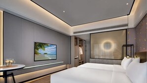 Un moderno cuarto de hotel con una cama grande, un televisor de pantalla plana en la pared, una mesa redonda pequeña con un florero y un ventanal con vista al mar.