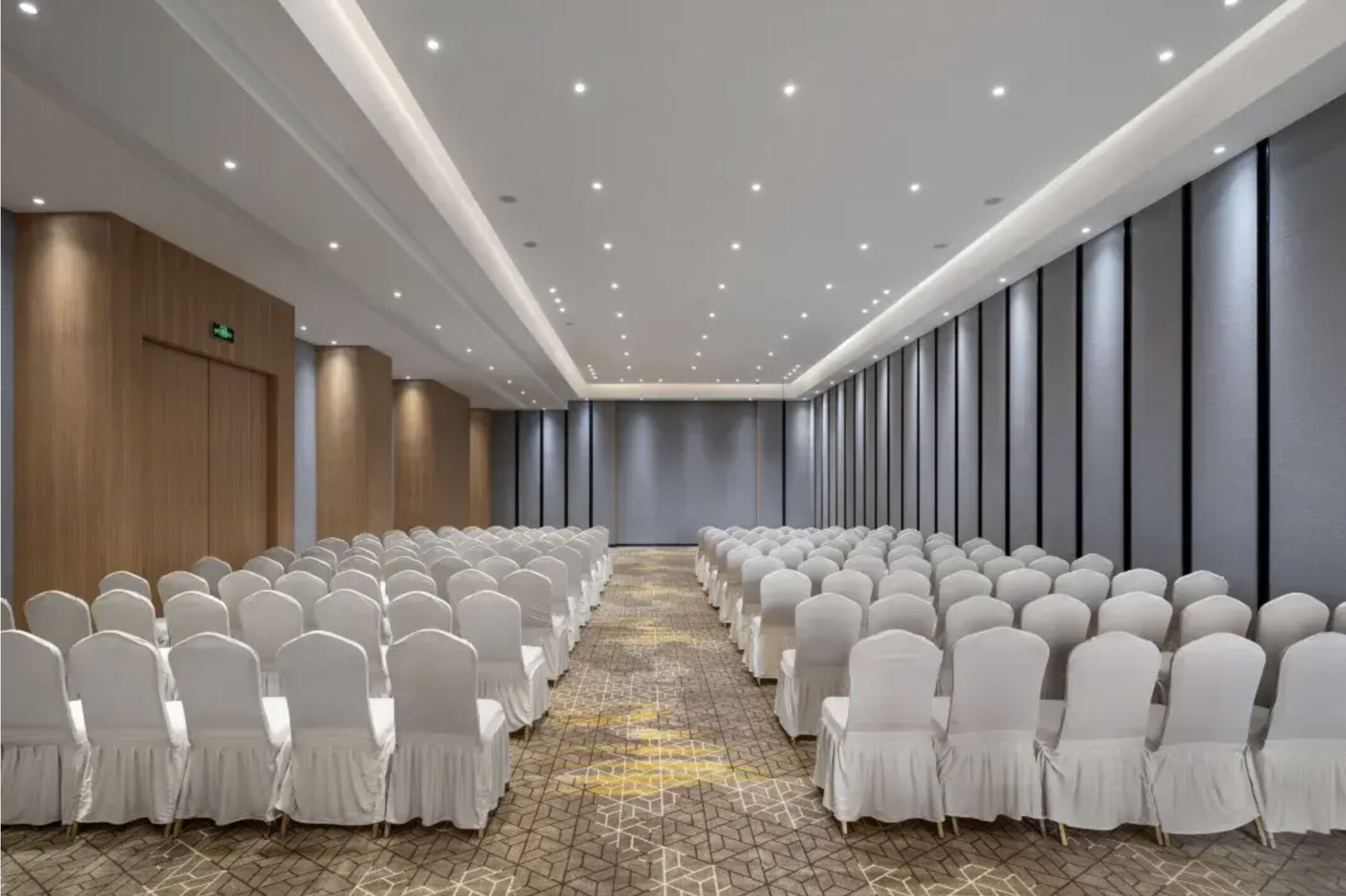 Un salón de conferencias con filas de sillas blancas, alfombra estampada y un diseño de techo moderno.