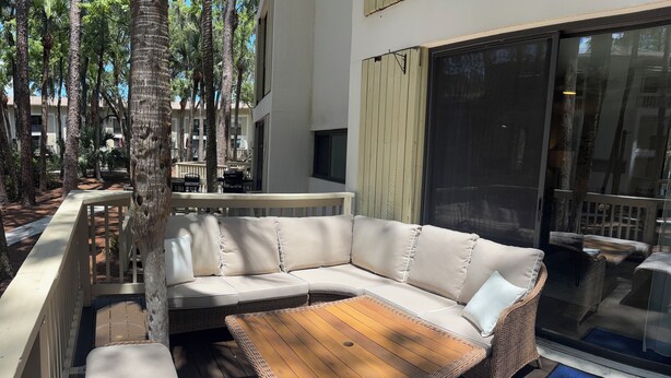 Ein moderner Außenbereich mit einer gebogenen Couch und einem Holztisch, umgeben von hohen Bäumen und einer Glastür zum Pool.