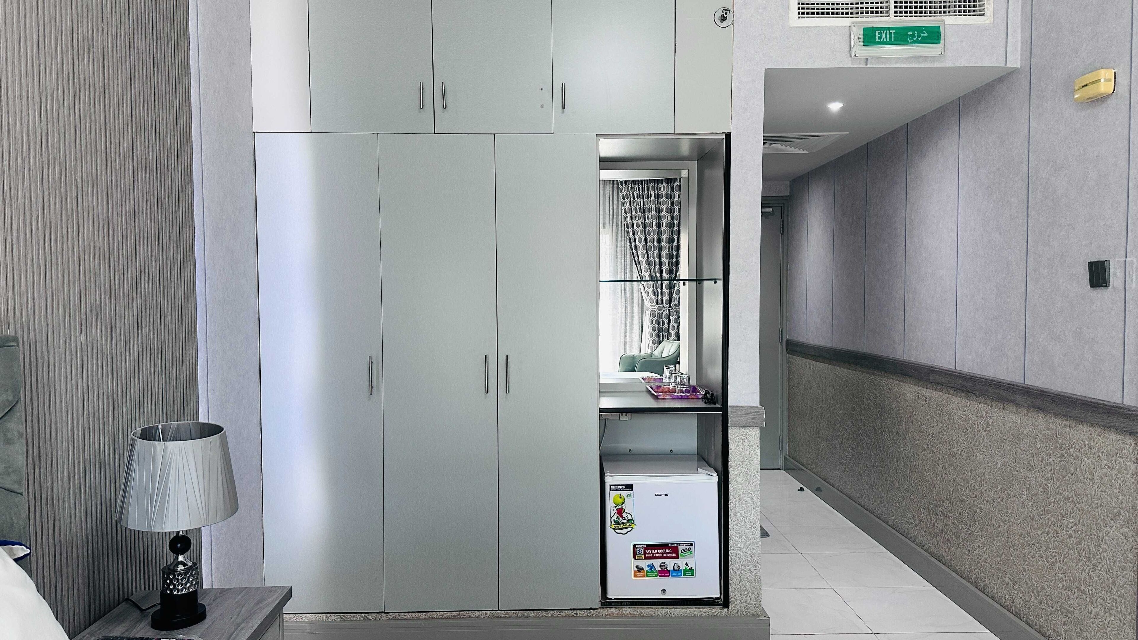 A hotel room with a wardrobe, a bedside table with a lamp, and a minibar.