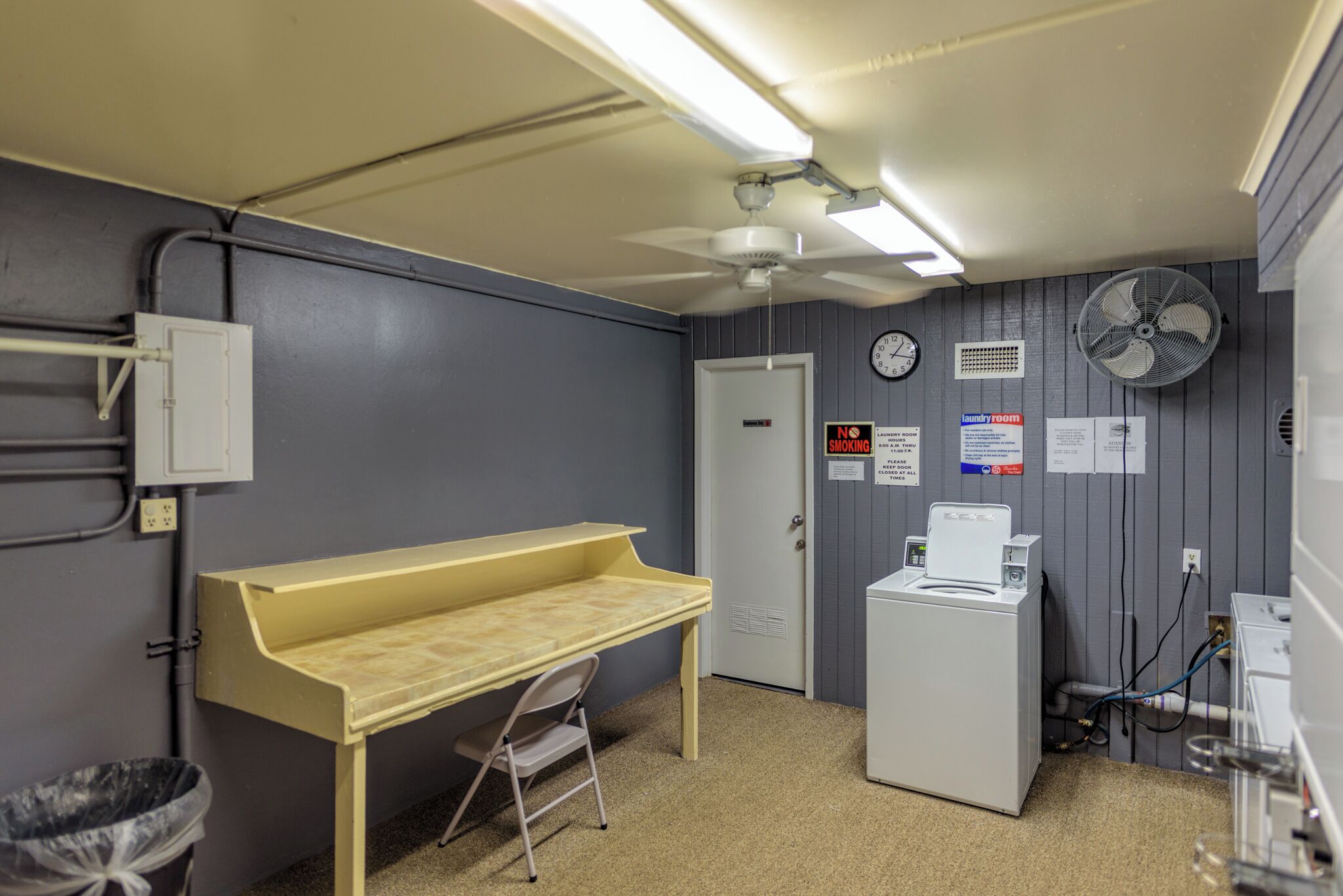 Une buanderie équipée d’un lave-linge, d’un sèche-linge, d’un bureau, d’une chaise, d’une poubelle et d’une horloge murale.