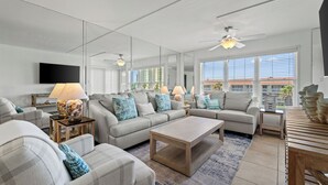 A modern living room with a large sofa set, a coffee table, and a TV mounted on the wall. There are multiple lamps and a ceiling fan.