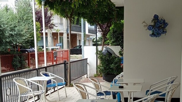 An outdoor patio area with tiled flooring, white metal chairs, and tables. There is a railing and greenery in the background. - albergo Atene (Riccione)