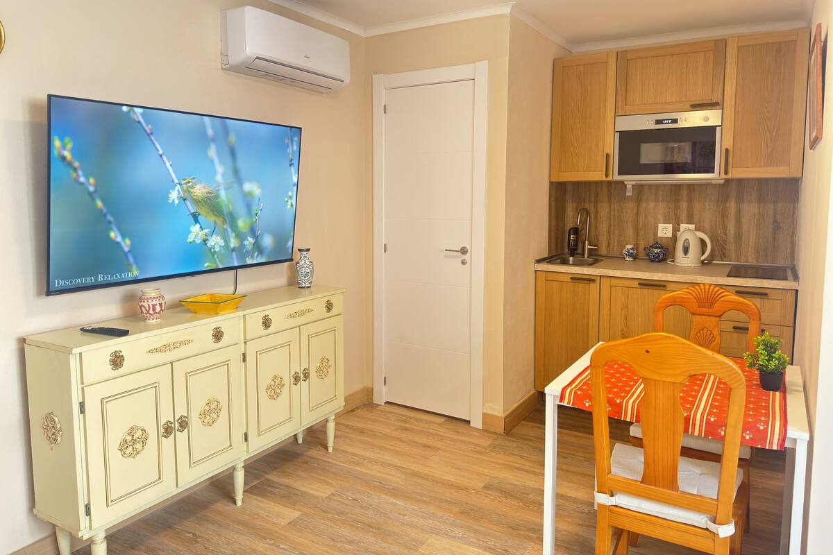 Een compacte woonruimte met een televisie, een eettafel, een keukenhoek met een magnetron en een kast.