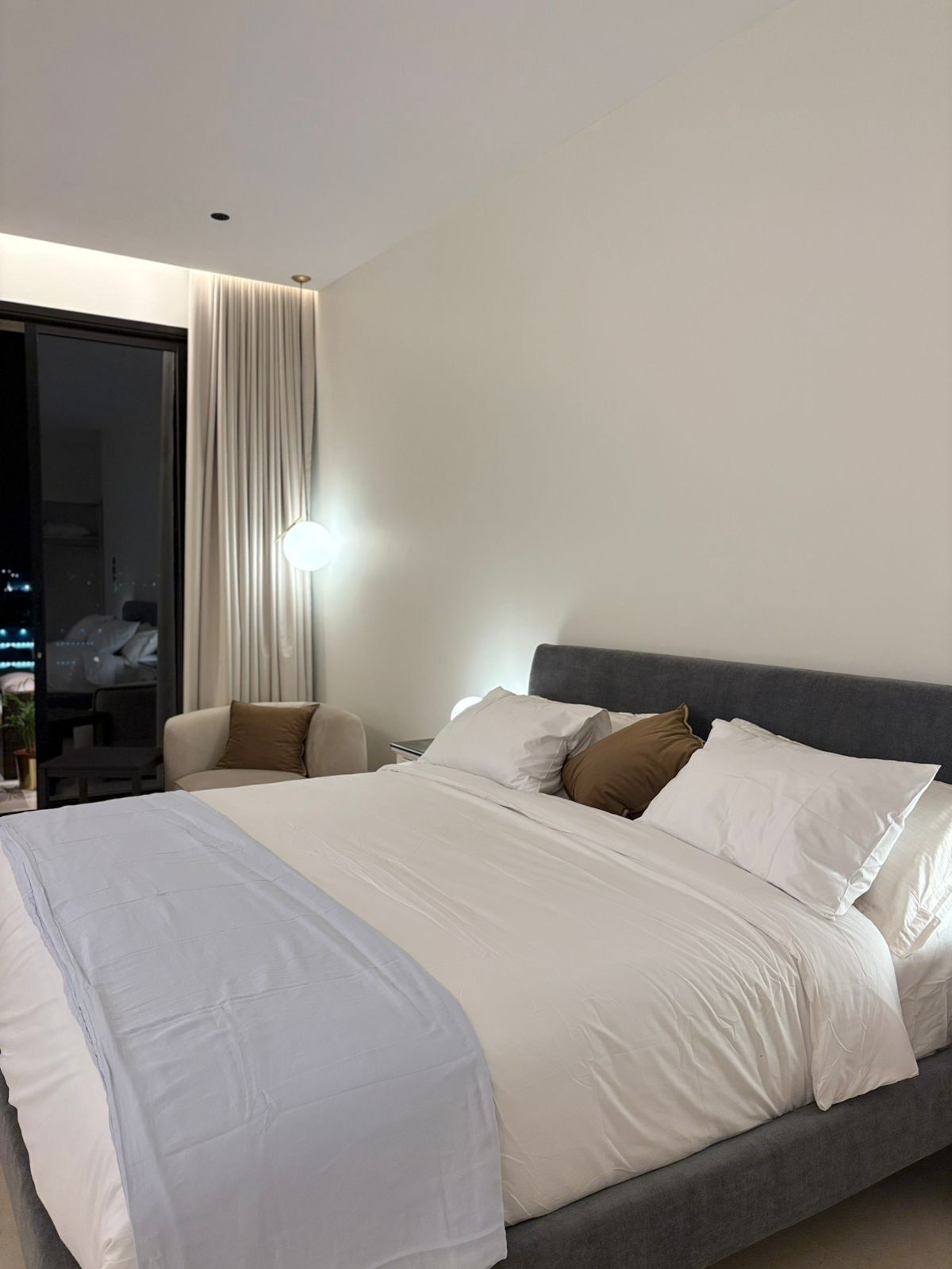 Quarto de hotel moderno com uma cama grande, um sofá, uma escrivaninha e vista para a cidade à noite.
