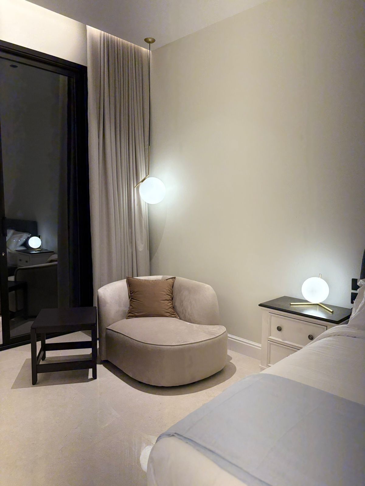 A modern hotel room with a curved sofa, a small table, a bedside table with a lamp, and a large bed.