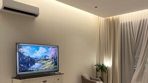 A modern bedroom with a flat-screen TV on a dresser, a bed with a white comforter, and a large window with sheer curtains. - Amazing studio with charming view in charming Taif (Taif)