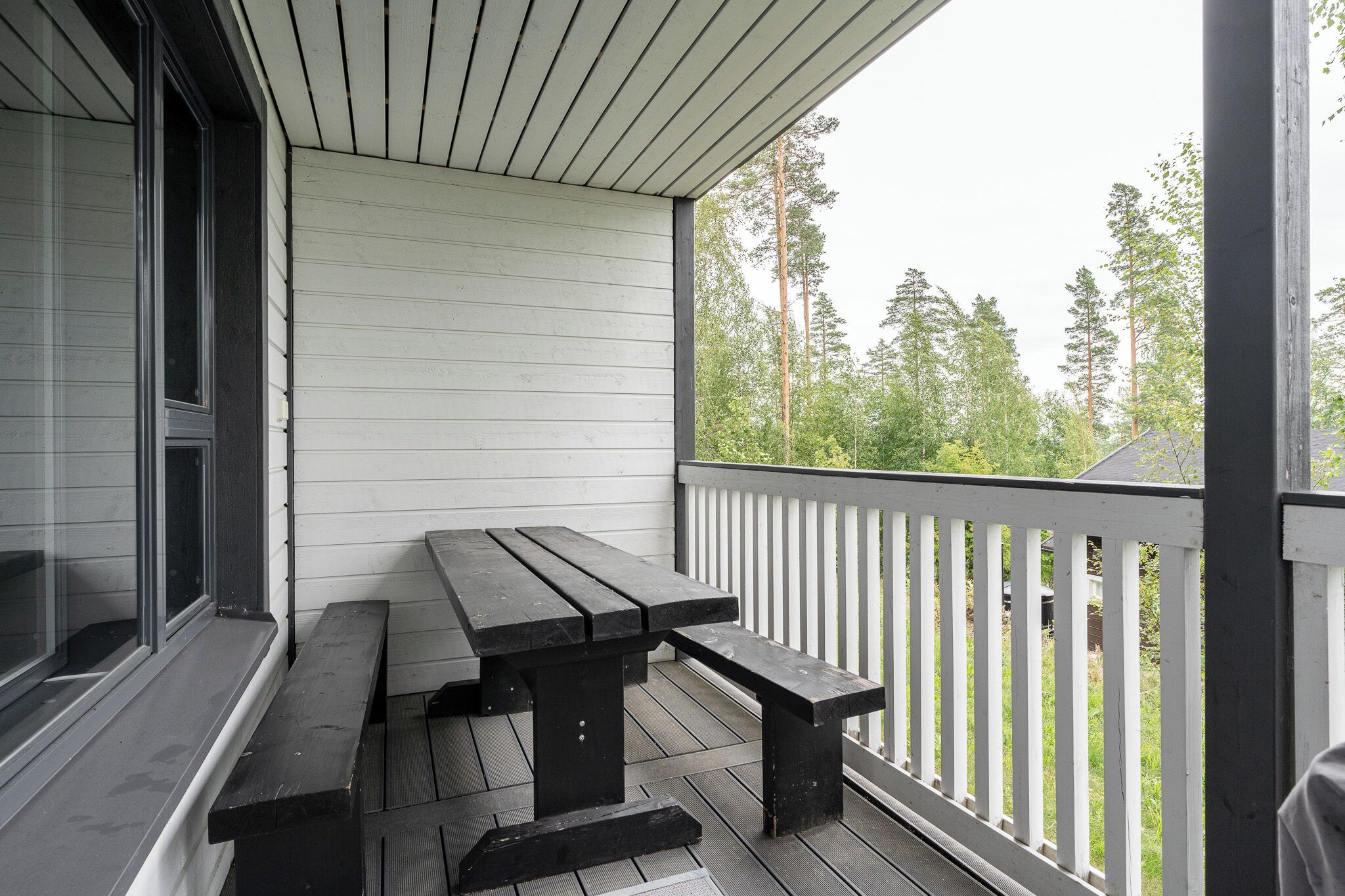 A wooden deck with a picnic table and benches, white railings, and a view of trees.