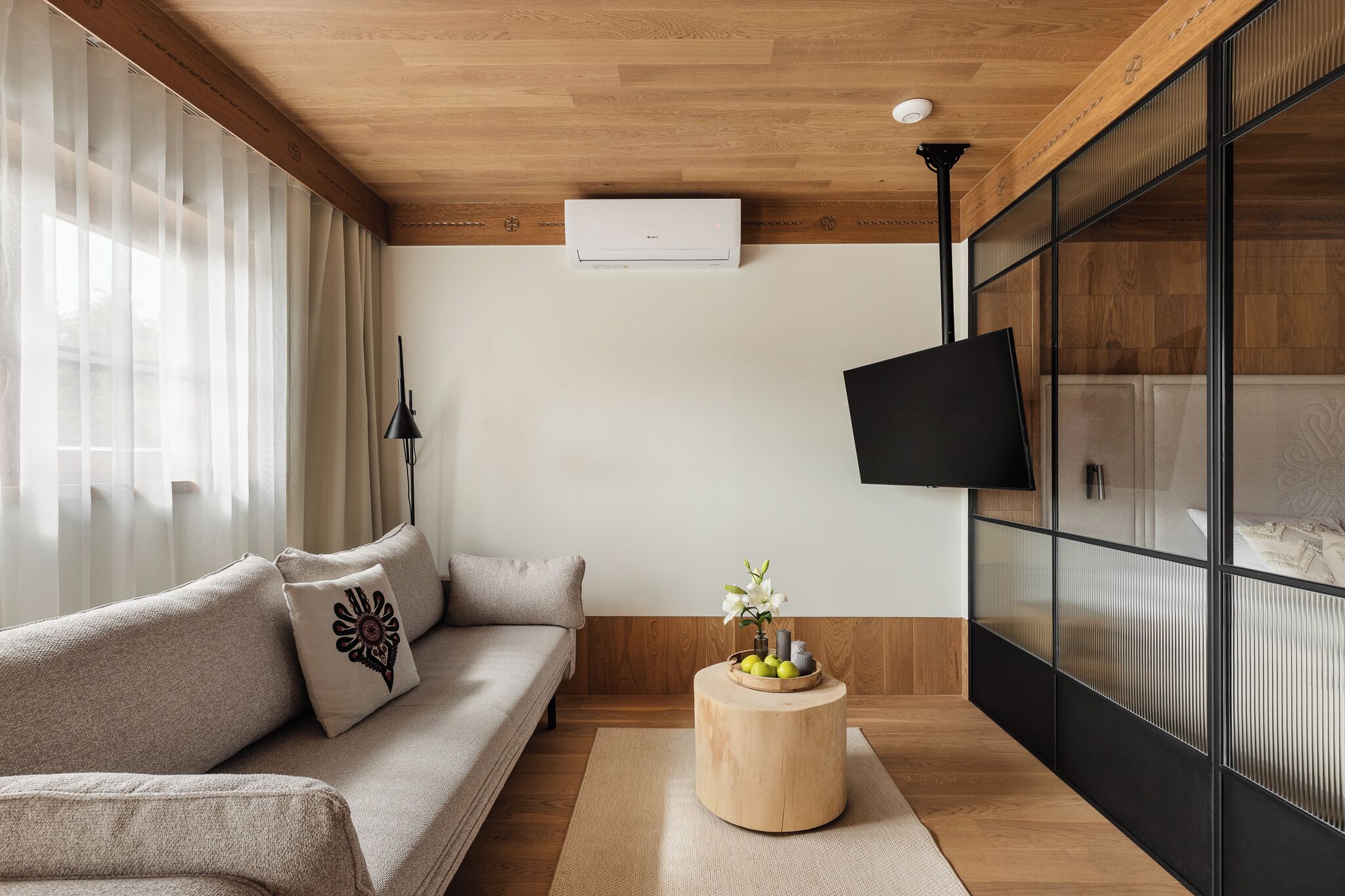 Una sala de estar moderna con techo de madera, un sofá color beige, una mesa de centro redonda de madera con objetos decorativos y un televisor de pantalla plana montado en una pared negra.
