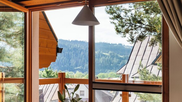 A round glass table with a white vase and a menu card, surrounded by wooden chairs, in a room with large windows offering a view of a forested area and a building with sloped roofs. - IJG Imperial Smrekowa (Zakopane)